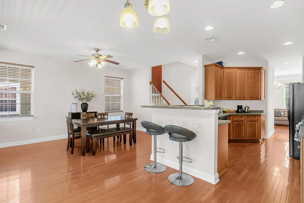 This image showcases a spacious dining table adjacent to the kitchen counter and bar stools, with a staircase in the background. The layout is ideal for family meals, casual gatherings, or entertaining friends.