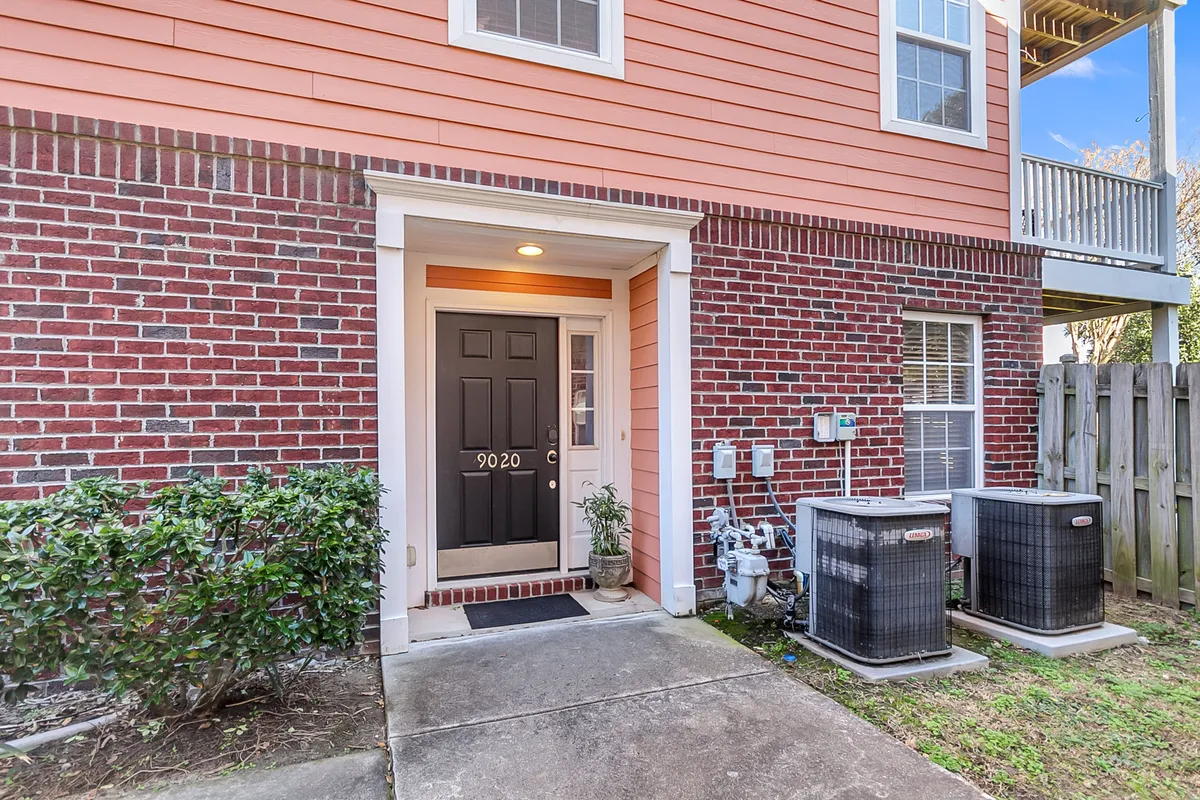 A welcoming front door framed by a brick facade, surrounded by manicured bushes and featuring visible air conditioning units. The approach is inviting, with a clear sense that this is a private entryway to a single-family home, suggesting a peaceful and self-contained setting for guests.