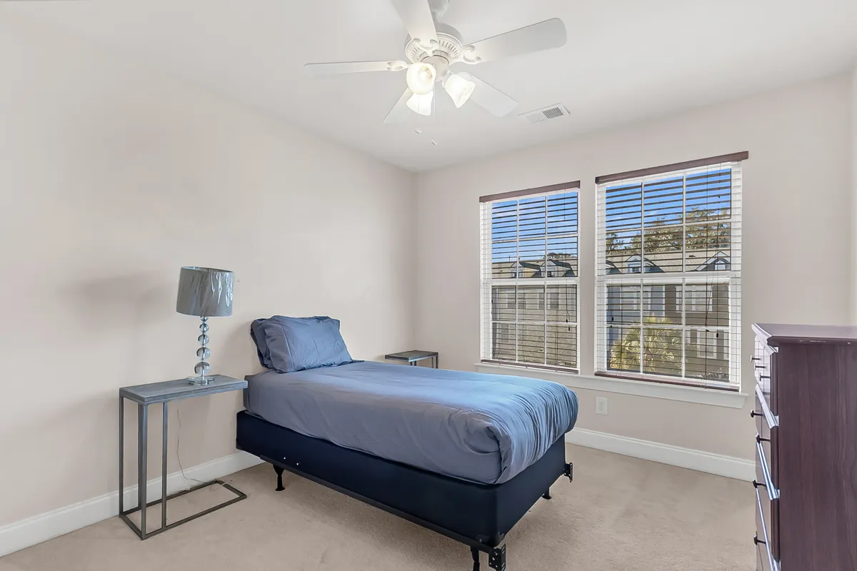 A tranquil bedroom with blue bedding, a soft lampshade table lamp, windows with blinds allowing in gentle light, and a chest of drawers. The space is styled simply, with an emphasis on comfort and a soothing vibe.