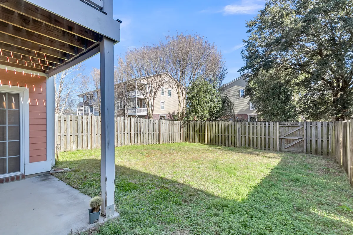 A fenced yard featuring a patio area with green grass and mature trees in the background. The inviting space suggests a safe environment for children to play, opportunities for gardening, and a tranquil setting for outdoor gatherings or quiet relaxation. Sunlight and open sky enhance the feeling of a serene outdoor retreat.