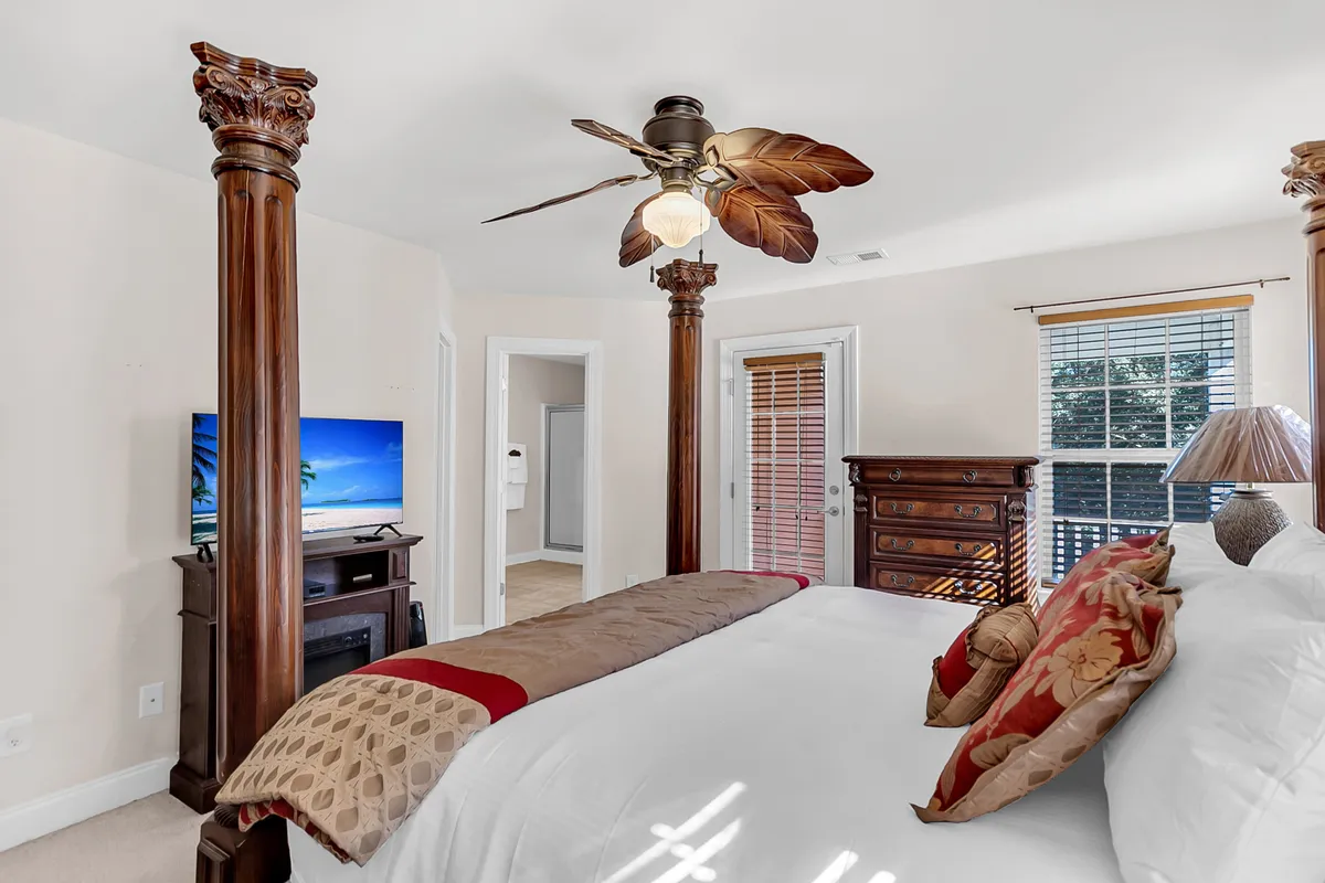 A spacious bedroom featuring a four-poster bed with plush bedding, a ceiling fan with a leaf design, and a television positioned for easy viewing. Natural light streams in, creating a cozy and inviting atmosphere ideal for relaxation.