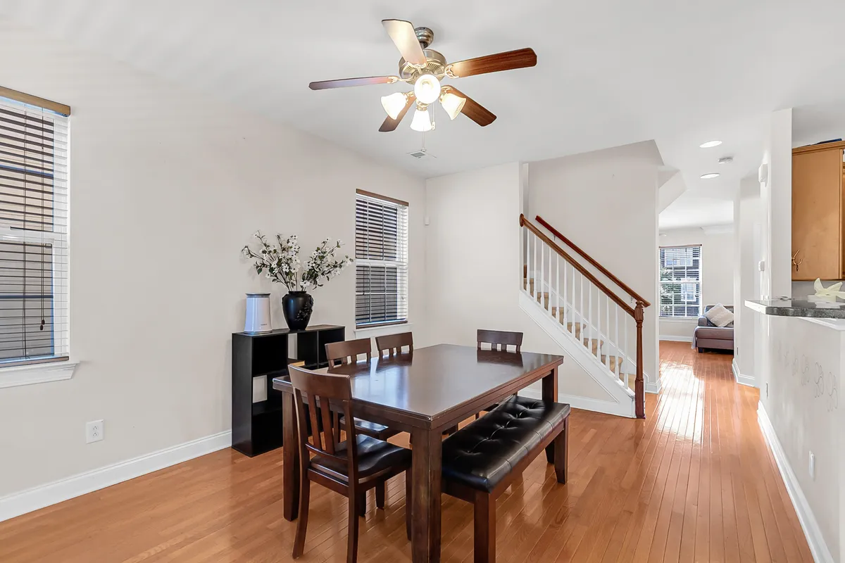 The image showcases a bright, welcoming dining area with a spacious table set under a ceiling fan, adjacent to a modern staircase. Natural light fills the space, creating a cozy atmosphere ideal for family gatherings, entertaining friends, or relaxing with a book by the window.