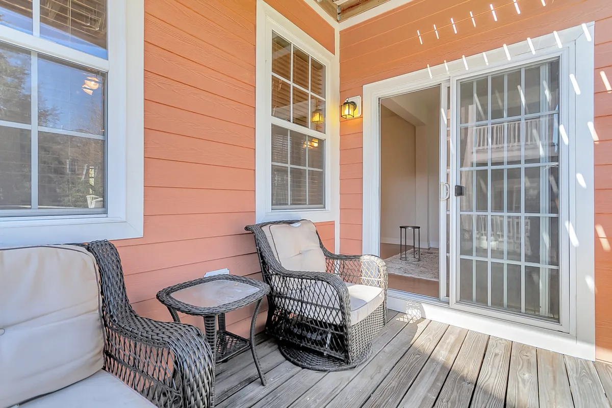 This image features a cozy patio setup with wicker chairs and a small table beside an orange wall and sliding glass door. The scene is bathed in gentle natural light, creating a welcoming atmosphere perfect for morning coffee, reading, or evening relaxation. The proximity to the indoors is emphasized by the sliding door, highlighting the flow between indoor comfort and outdoor enjoyment.