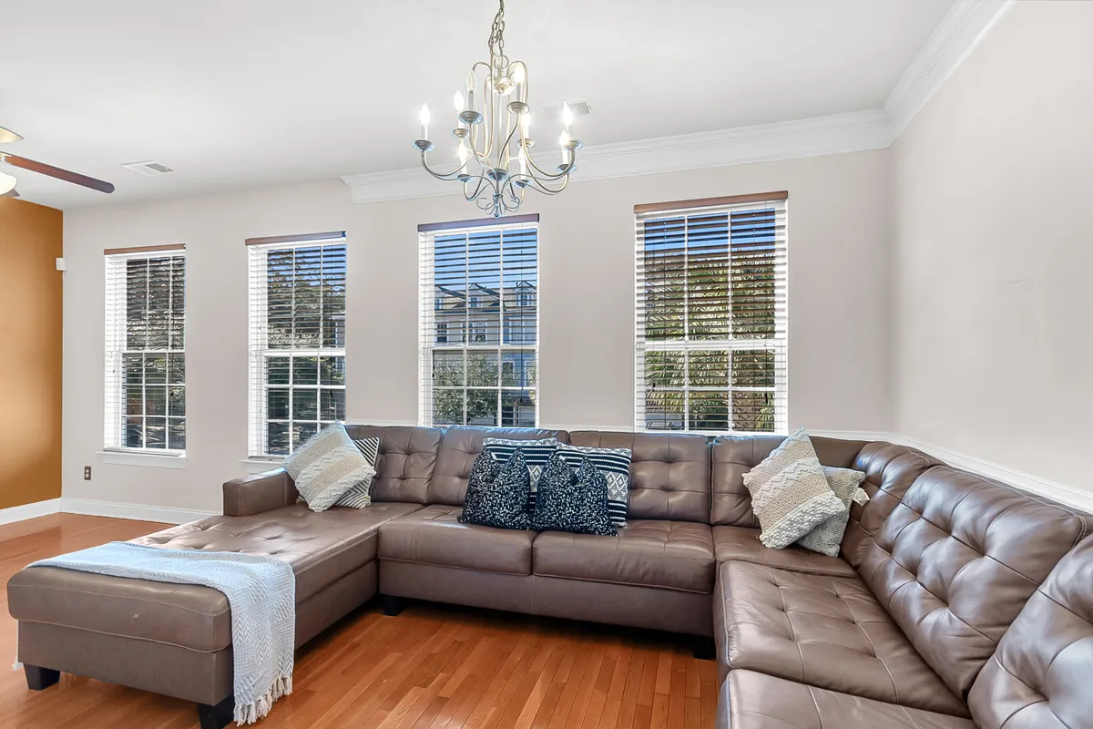 A bright, open living room featuring a large leather sectional sofa arranged around a central coffee table. Sunlight pours in from multiple windows, highlighting a stylish chandelier above. The inviting space is clearly designed for relaxation and group gatherings, with ample seating and a cozy, welcoming ambiance.