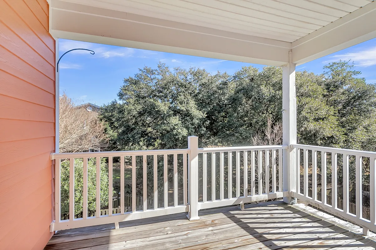 A wooden patio railing overlooks lush green trees beneath a clear blue sky. The scene suggests peaceful solitude or shared moments, such as sipping coffee at sunrise, reading a book, or simply enjoying the sounds of nature. The view emphasizes the patio’s connection to its natural surroundings.